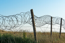 Barbed Wire Fence Against A Blue Sky And A Hilly Grassy Area. Closed Military Area