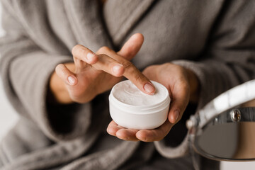 Hand moisturizer cream close-up. Hands cream for protect skin from dryness in bedroom. African american woman in bathrobe with hands moisturizing cream doing morning beauty routine.