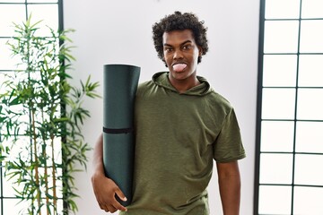 Poster - African man with curly hair holding yoga mat at studio sticking tongue out happy with funny expression. emotion concept.