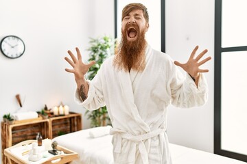 Wall Mural - Redhead man with long beard wearing bathrobe at wellness spa crazy and mad shouting and yelling with aggressive expression and arms raised. frustration concept.