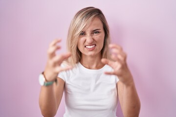 Canvas Print - Young blonde woman standing over pink background shouting frustrated with rage, hands trying to strangle, yelling mad