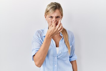 Canvas Print - Beautiful blonde woman standing over white background smelling something stinky and disgusting, intolerable smell, holding breath with fingers on nose. bad smell