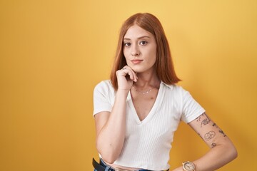 Poster - Young redhead woman standing over yellow background with hand on chin thinking about question, pensive expression. smiling with thoughtful face. doubt concept.