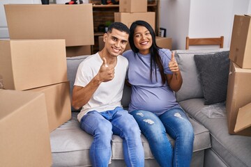 Poster - Young hispanic couple expecting a baby sitting on the sofa at new home smiling happy and positive, thumb up doing excellent and approval sign