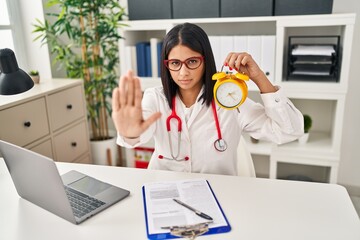 Wall Mural - Young hispanic doctor woman holding alarm clock at the clinic with open hand doing stop sign with serious and confident expression, defense gesture