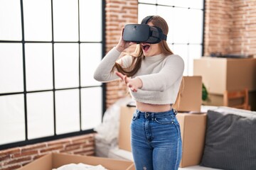 Poster - Young blonde woman using virtual reality goggles sitting on sofa at new home