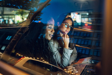 two multiracial business woman riding in a car and using a laptop