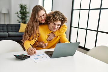 Poster - Young caucasian couple hugging each other doing family accounting using laptop at home