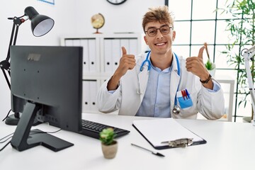 Sticker - Young caucasian doctor man working at the clinic success sign doing positive gesture with hand, thumbs up smiling and happy. cheerful expression and winner gesture.