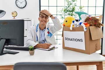 Poster - Young caucasian doctor man working at the clinic with toys donations box for children stressed and frustrated with hand on head, surprised and angry face