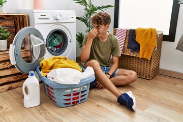 Poster - Young caucasian man putting dirty laundry into washing machine tired rubbing nose and eyes feeling fatigue and headache. stress and frustration concept.