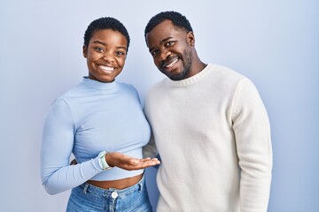 Sticker - Young african american couple standing over blue background pointing aside with hands open palms showing copy space, presenting advertisement smiling excited happy