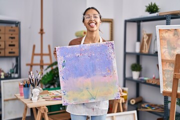 Sticker - Young brazilian woman holding painter canvas at art studio sticking tongue out happy with funny expression.