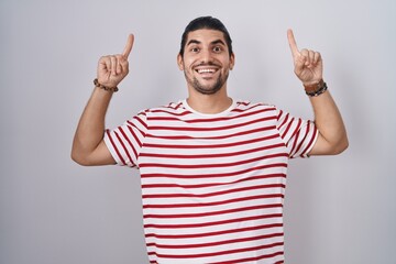 Wall Mural - Hispanic man with long hair standing over isolated background smiling amazed and surprised and pointing up with fingers and raised arms.