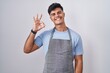 © Krakenimages.com - Hispanic young man wearing apron over white background smiling positive doing ok sign with hand and fingers. successful expression.