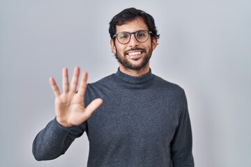 Sticker - Handsome latin man standing over isolated background showing and pointing up with fingers number five while smiling confident and happy.