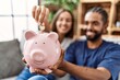 © Krakenimages.com - Man and woman couple smiling confident insert coin on piggy bank at home