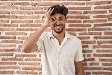 Poster - Arab man with beard standing over bricks wall background doing ok gesture with hand smiling, eye looking through fingers with happy face.