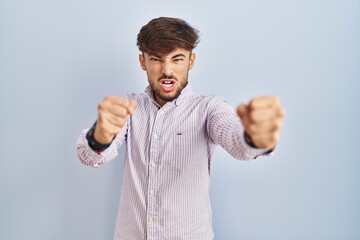 Wall Mural - Arab man with beard standing over blue background angry and mad raising fists frustrated and furious while shouting with anger. rage and aggressive concept.