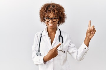 Canvas Print - Young african american woman wearing doctor uniform and stethoscope smiling and looking at the camera pointing with two hands and fingers to the side.