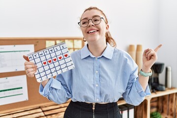 Wall Mural - Young redhead woman holding heart calendar at the office smiling happy pointing with hand and finger to the side