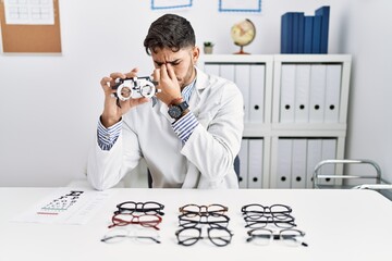 Poster - Young optician man holding optometry glasses tired rubbing nose and eyes feeling fatigue and headache. stress and frustration concept.