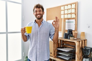 Wall Mural - Young handsome man drinking a cup coffee at the office doing ok sign with fingers, smiling friendly gesturing excellent symbol