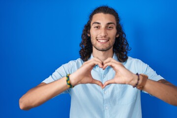 Poster - Young hispanic man standing over blue background smiling in love doing heart symbol shape with hands. romantic concept.