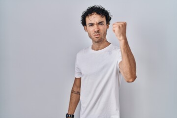 Wall Mural - Hispanic man standing over isolated background angry and mad raising fist frustrated and furious while shouting with anger. rage and aggressive concept.