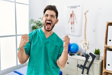 Sticker - Young man with beard working at pain recovery clinic very happy and excited doing winner gesture with arms raised, smiling and screaming for success. celebration concept.