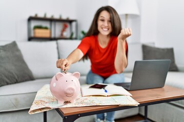 Poster - Young brunette woman putting euro coin in piggy bank saving for travel screaming proud, celebrating victory and success very excited with raised arm