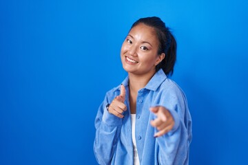 Wall Mural - Asian young woman standing over blue background pointing fingers to camera with happy and funny face. good energy and vibes.