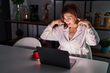 Poster - Middle age hispanic woman using laptop at home at night covering ears with fingers with annoyed expression for the noise of loud music. deaf concept.