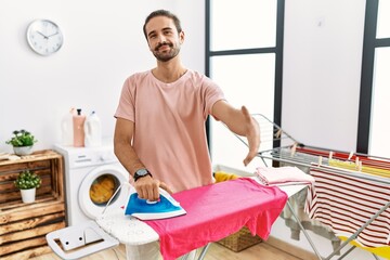 Canvas Print - Young hispanic man ironing clothes at home smiling friendly offering handshake as greeting and welcoming. successful business.