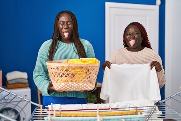 Two african women hanging clothes at clothesline sticking tongue out happy with funny expression.