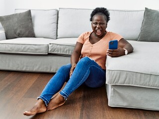 Poster - Young african woman using smartphone sitting on the floor at home angry and mad screaming frustrated and furious, shouting with anger. rage and aggressive concept.