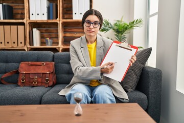 Poster - Young hispanic psychiatrist woman working at consultation office with clipboard relaxed with serious expression on face. simple and natural looking at the camera.