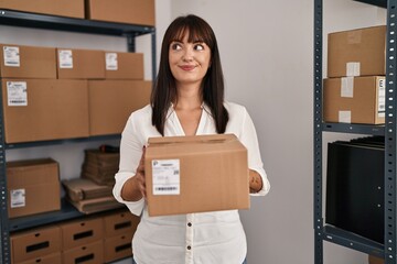 Wall Mural - Young brunette woman working at small business ecommerce holding package smiling looking to the side and staring away thinking.