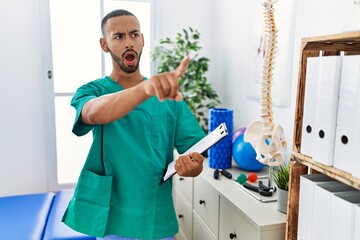 Wall Mural - African american physiotherapist man working at pain recovery clinic pointing with finger surprised ahead, open mouth amazed expression, something on the front