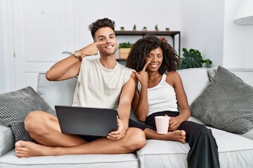 Wall Mural - Young interracial couple using laptop at home sitting on the sofa pointing with hand finger to face and nose, smiling cheerful. beauty concept