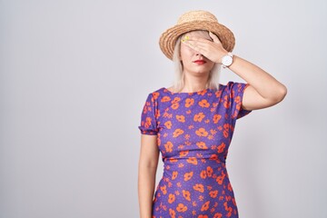 Poster - Young caucasian woman wearing flowers dress and summer hat covering eyes with hand, looking serious and sad. sightless, hiding and rejection concept