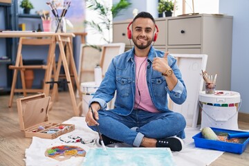 Sticker - Young hispanic man painter sitting on the floor at art studio smiling happy and positive, thumb up doing excellent and approval sign