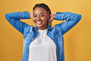 Poster - African american woman with braids standing over yellow background relaxing and stretching, arms and hands behind head and neck smiling happy