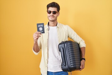 Canvas Print - Young hispanic man holding suitcase going on summer vacation holding italian passport smiling looking to the side and staring away thinking.