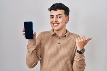 Wall Mural - Non binary person holding smartphone showing blank screen pointing thumb up to the side smiling happy with open mouth