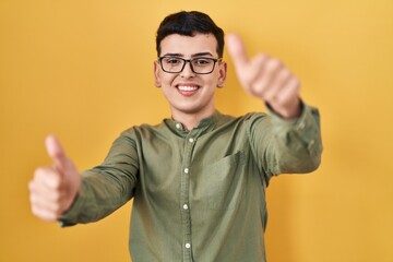 Poster - Non binary person standing over yellow background approving doing positive gesture with hand, thumbs up smiling and happy for success. winner gesture.