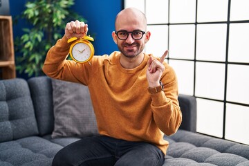 Wall Mural - Young bald man with beard holding alarm clock smiling happy pointing with hand and finger to the side