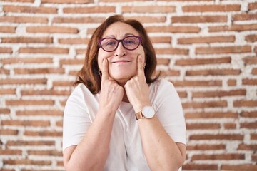 Poster - Senior woman with glasses standing over bricks wall smiling with open mouth, fingers pointing and forcing cheerful smile