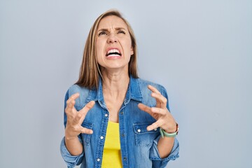 Poster - Young blonde woman standing over blue background crazy and mad shouting and yelling with aggressive expression and arms raised. frustration concept.