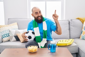 Canvas Print - Young hispanic man with beard and tattoos football hooligan holding ball supporting team smiling happy pointing with hand and finger to the side